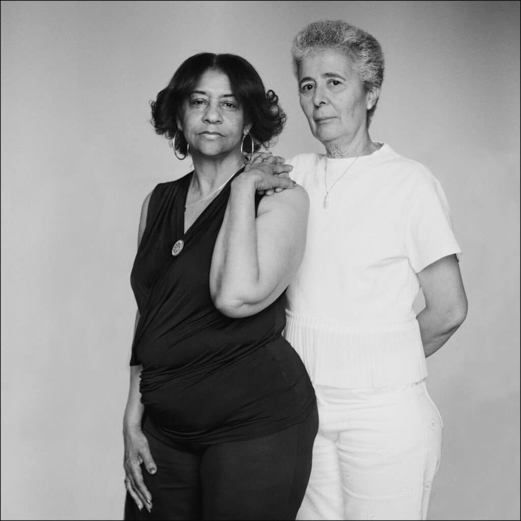 Dos mujeres posan de pie, una con el brazo apoyado sobre el hombro de la otra. La mujer de la izquierda viste de negro y la de la derecha de blanco, ambas miran con serenidad hacia la cámara.
