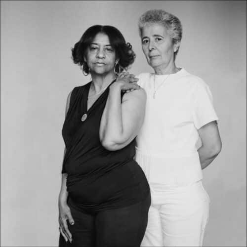 Dos mujeres posan de pie, una con el brazo apoyado sobre el hombro de la otra. La mujer de la izquierda viste de negro y la de la derecha de blanco, ambas miran con serenidad hacia la cámara.