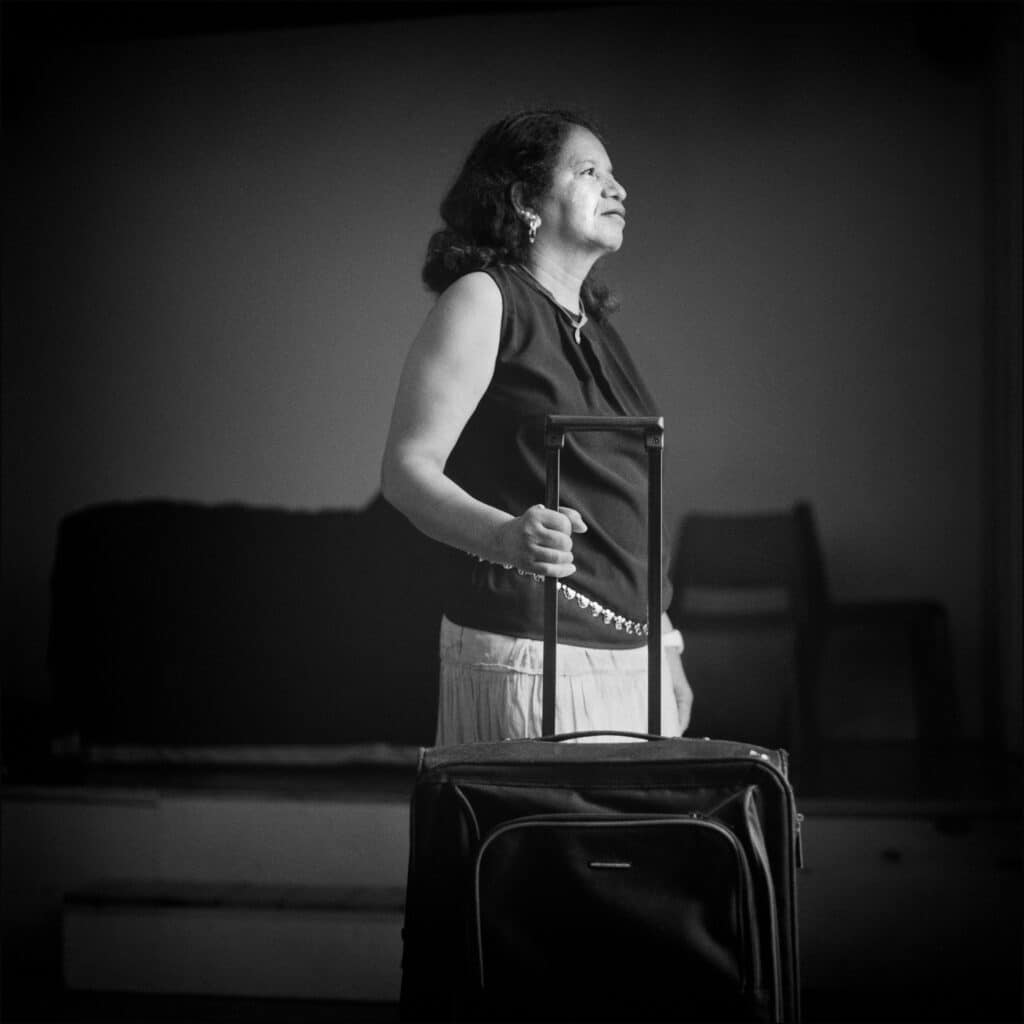 Mujer de pie sosteniendo el asa de una maleta, mirando hacia arriba con expresión serena.