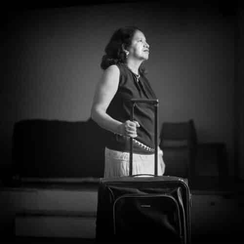 Mujer de pie sosteniendo el asa de una maleta, mirando hacia arriba con expresión serena.