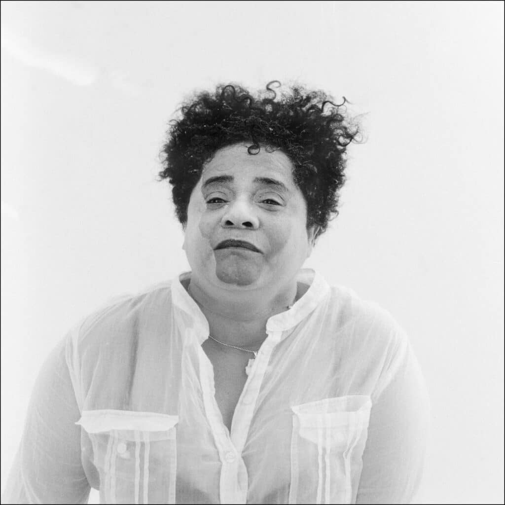 Mujer con el cabello rizado mirando al frente, vestida con una camisa blanca translúcida.