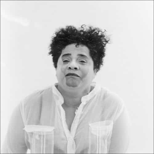 Mujer con el cabello rizado mirando al frente, vestida con una camisa blanca translúcida.