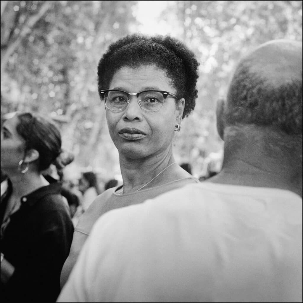 Mujer con gafas mirando a cámara entre un grupo de personas en un espacio al aire libre.